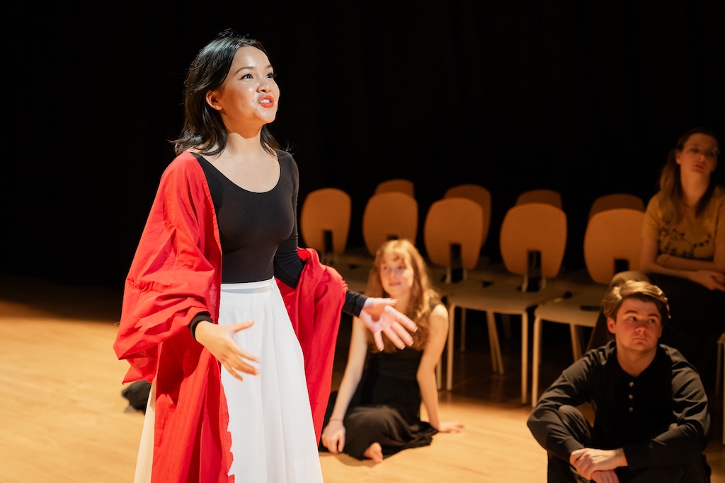 An actor wearing a bright red cape stands on stage in mid-performance while two other actors sit near her on stage.